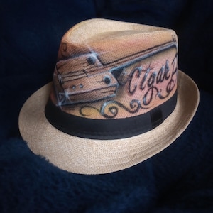 May include: A tan fedora hat with a black band and the words "Cigar Bar" painted on the side. The hat features a detailed illustration of a cigar box. The hat is made of woven material.