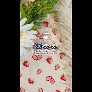 May include: A sticker sheet with three cartoon characters and the text "Start Digging In Your Butt" is displayed on a light pink fabric with a strawberry pattern. A white flower and green foliage are visible on the left side of the image.
