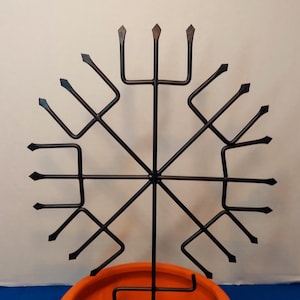 May include: A black metal decorative stand with a circular design, featuring a central vertical support and radiating arms with pointed ends. It sits atop an orange, round, ceramic bowl. The stand's design is symmetrical and geometric.