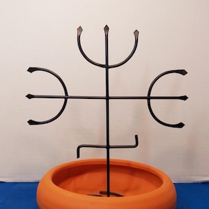 May include: An orange ceramic planter with a black metal decorative plant stand. The stand has a unique design with curved and pointed elements. The planter is round and sits on a blue surface.