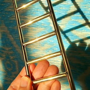 Może przedstawiać: Mała, złota drabina z sześcioma szczeblami. Drabina trzymana jest w dłoni na tle niebiesko-białego wzoru. Drabina wykonana jest z metalu i ma błyszczące wykończenie. Przedmiot jest prawdopodobnie elementem dekoracyjnym lub miniaturowym akcesorium.