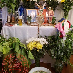May include: A table set for a spiritual ritual, adorned with fresh flowers, candles, and religious figurines. Various bottles and glasses are arranged on a white tablecloth, with a rosary and a bowl of petals. The logo "Ritual Scent" is visible.