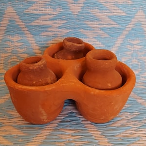 May include: A terracotta clay holder with three small terracotta pots. The holder has a unique shape with three rounded openings for the pots.