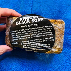 May include: A bar of African Black Soap, wrapped in clear plastic, with a black label. The label reads "AFRICAN BLACK SOAP 100% NATURAL" and provides usage instructions. The soap is made in West Africa and weighs 8 oz.