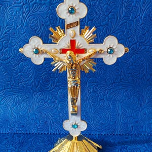 Könnte beinhalten: Ein dekoratives Kruzifix mit einer goldfarbenen Jesusfigur, einem roten Kreuz und weißen Kreuzarmen. Das Kreuz hat goldfarbene Akzente, blaue Edelsteine und einen goldenen Sockel. Der Hintergrund ist eine strukturierte blaue Oberfläche.