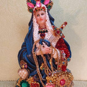May include: A blue and gold figurine of the Virgin Mary, adorned with colourful jewels and beads. The figurine is wearing a crown and a blue robe with a white veil. The base of the figurine is decorated with gold and red jewels.