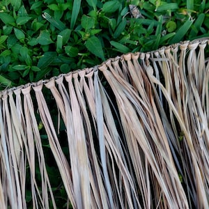 Może przedstawiać: Zbliżenie naturalnego, tkanego materiału strzechy z długimi, wysuszonymi, beżowymi liśćmi. Strzecha jest przymocowana do tkanej krawędzi. Tło to zielona trawa.