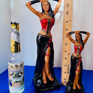 May include: Two statues of a woman in a red corset and black skirt, with arms raised above her head. A bottle of Florida Water cologne is on the left. The statues are approximately 10 inches and 6 inches tall, respectively.