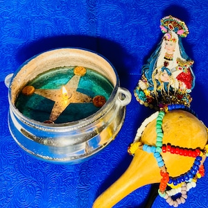 May include: A silver-toned metal bowl containing a lit candle with a star-shaped wick holder, a small figurine of a person wearing a blue and red robe with ornate headwear, and a tan gourd rattle adorned with colorful beads in red, blue, green, and white.
