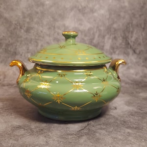 Może przedstawiać: Zielona ceramiczna miska z pokrywką i złotymi akcentami. Naczynie ma wzór kratki ze złotymi wzorami gwiazd. Uchwyty i obręcz są wykończone złotem. Pokrywka ma mały uchwyt i jest również ozdobiona złotem.