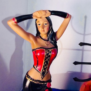 May include: A figurine of a woman with dark hair and a red corset-style top with black lacing and gold trim. She is wearing a black skirt with gold trim and red roses. Her arms are raised above her head.