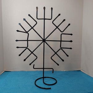 May include: Black metal wall decor featuring a symmetrical design with a central hub and radiating arms. The arms terminate in stylized arrowheads or fork-like shapes. The base has a circular design. The piece is displayed against a blue surface.