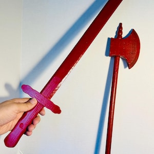 May include: A red wooden sword and axe set. The sword has a cross-shaped handle and a long, straight blade. The axe features a rectangular head and a long handle. Both items are painted a solid, vibrant red color.