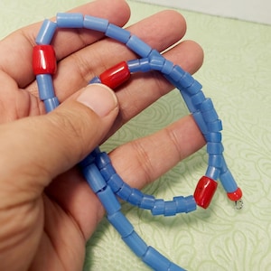 Może przedstawiać: Niebieski naszyjnik z koralików z czerwonymi akcentami w kształcie beczki. Naszyjnik składa się z małych, cylindrycznych niebieskich koralików przeplatanych większymi, czerwonymi koralikami. Naszyjnik jest trzymany w dłoni na jasnym zielonym tle.