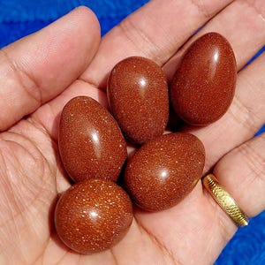 Może przedstawiać: Kolekcja sześciu wypolerowanych kamieni w kształcie jajka w odcieniach brązu i złota. Kamienie mają nakrapiany, błyszczący wygląd i są trzymane w dłoni. Złoty pierścionek jest widoczny na palcu.