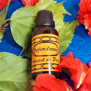 May include: A small brown glass bottle with a black cap and a yellow label. The label reads "Parfum d'amour Aimant de Paris" in black script. The bottle is surrounded by green leaves and red flowers.