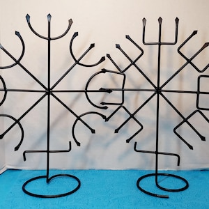May include: Two black metal decorative wall art pieces. Each piece features a symmetrical design with curved and pointed elements radiating from a central point. The pieces stand on circular bases and are suitable for wall decor.