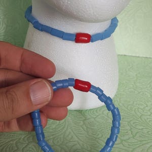 Może przedstawiać: Jasnoniebieski zestaw naszyjnika i bransoletki z koralików. Naszyjnik jest wokół białej głowy manekina. Bransoletka jest trzymana w dłoni. Oba elementy mają czerwone prostokątne akcenty. Koraliki są małe i kwadratowe.