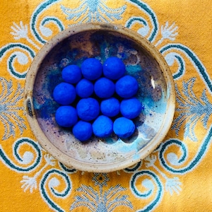May include: A ceramic bowl with a blue and brown glaze holds a collection of vibrant blue spheres. The bowl sits on a yellow textile with a teal and white floral pattern, creating a striking contrast of colors and textures.