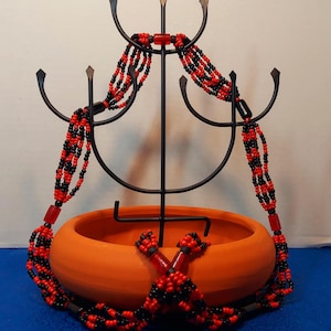 May include: A black metal jewelry display stand holding multiple strands of red and black beaded necklaces. The necklaces are draped over the stand and rest in an orange ceramic bowl. The background is a blue surface.