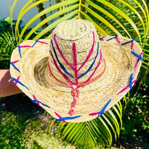 May include: A straw hat with a wide brim and a rounded crown. The hat is decorated with pink and blue stitching, creating a colorful pattern. The hat is made of woven straw and has a natural, light brown color.