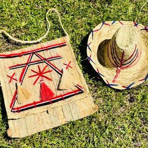 Puede incluir: Un bolso de hombro de paja con detalles bordados en rojo, negro y blanco y borlas, junto a un sombrero de paja con ribetes rojos y azules. El bolso tiene un diseño tejido y una correa larga.