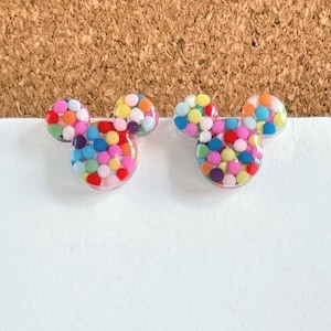 May include: A pair of colorful Mickey Mouse-shaped earrings with a confetti design. The earrings are made of clear resin and filled with small, colorful dots.