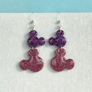 K&ouml;nnte beinhalten: Ein Paar Ohrringe mit zwei &uuml;bereinanderliegenden glitzernden Mickey-Maus-K&ouml;pfen. Die obere Schicht ist aus lila Glitzer und die untere Schicht aus rosa Glitzer.