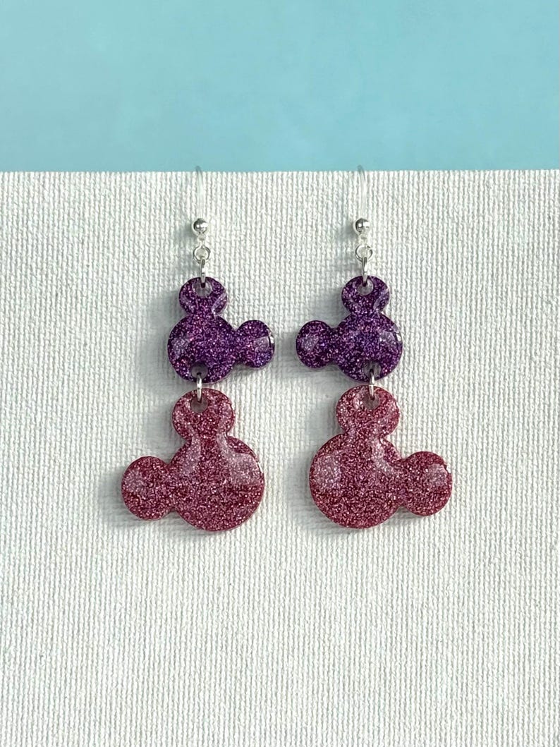 K&ouml;nnte beinhalten: Ein Paar Ohrringe mit zwei &uuml;bereinanderliegenden glitzernden Mickey-Maus-K&ouml;pfen. Die obere Schicht ist aus lila Glitzer und die untere Schicht aus rosa Glitzer.