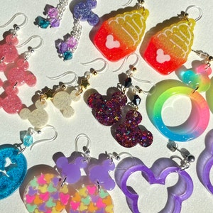 May include: Assortment of colorful resin earrings featuring various designs, including Mickey Mouse ears, ice cream cones, and rainbow swirls. The earrings are handmade and have a glittery finish.