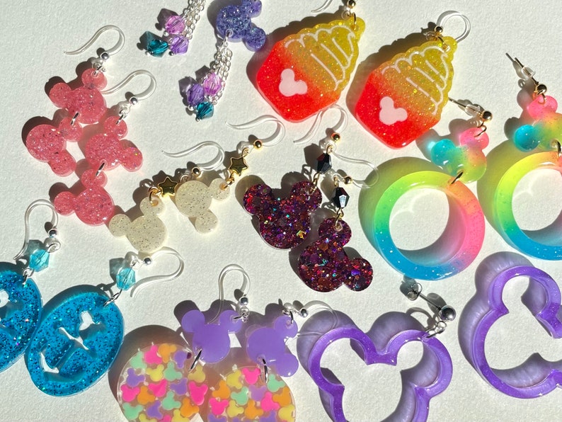 May include: Assortment of colorful resin earrings featuring various designs, including Mickey Mouse ears, ice cream cones, and rainbow swirls. The earrings are handmade and have a glittery finish.