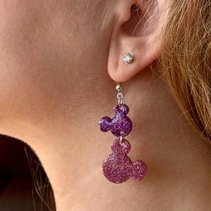 K&ouml;nnte beinhalten: Lila und rosa glitzernde Mickey Mouse Ohrh&auml;nger. Die Ohrringe sind silberfarben und haben einen kleinen, klaren Strassstein im Ohr.