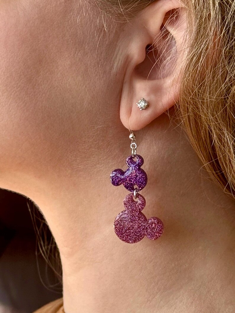 K&ouml;nnte beinhalten: Lila und rosa glitzernde Mickey Mouse Ohrh&auml;nger. Die Ohrringe sind silberfarben und haben einen kleinen, klaren Strassstein im Ohr.