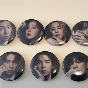 Op de afbeelding: Een set van zeven ronde buttons, elk voorzien van een afzonderlijk zwart-wit portret van een ander persoon. De glanzende buttons zijn gerangschikt in twee rijen en tonen close-up portretten van zeven verschillende individuen.