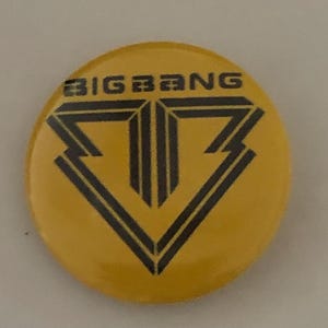 Puede incluir: Una chapa redonda de color amarillo con un logotipo gráfico negro de una letra B estilizada y el texto BIGBANG impreso en letras mayúsculas negras en la parte superior de la superficie de la chapa. El diseño está centrado sobre el fondo amarillo.