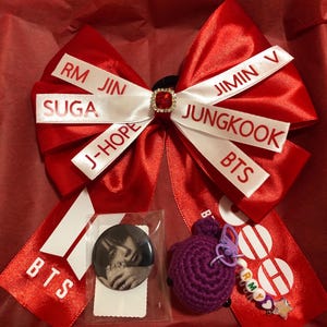 Pode incluir: Um grande laço de cabelo de cetim vermelho com fitas brancas impressas com os nomes RM, JIN, SUGA, J-HOPE, JIMIN, V, JUNGKOOK e BTS, centrado com um strass vermelho. Inclui um botão fotográfico circular e um pequeno pendente de crochê roxo.