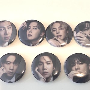 May include: A set of seven circular pin-back badges, each featuring a distinct black and white portrait of a different individual. The portraits show close-up views of faces with various hand placements near the chin or cheek, arranged in two rows.