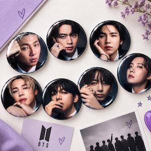 Puede incluir: Siete chapas circulares con retratos individuales de los miembros del grupo musical BTS. Cada chapa muestra una fotografía en primer plano de un miembro con traje oscuro. Se incluye una tarjeta morada con el logotipo de BTS.