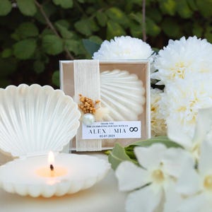 Può includere: Una candela bianca a forma di conchiglia è accesa, accanto a una scatola bianca a forma di conchiglia contenente un oggetto simile. La scatola è decorata con un nastro, fiori secchi e una perla. Il testo sulla scatola recita "THANK YOU FOR CELEBRATING OUR DAY WITH US ALEX MIA 02.07.2025 ∞". Fiori bianchi sullo sfondo.