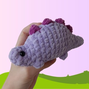 Puede incluir: Un juguete de peluche de dinosaurio de crochet morado con detalles en rosa. El dinosaurio tiene una cola larga y una cabeza peque&ntilde;a y redonda con ojos negros.