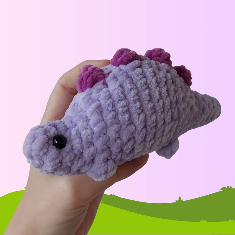 Puede incluir: Un juguete de peluche de dinosaurio de crochet morado con detalles en rosa. El dinosaurio tiene una cola larga y una cabeza peque&ntilde;a y redonda con ojos negros.