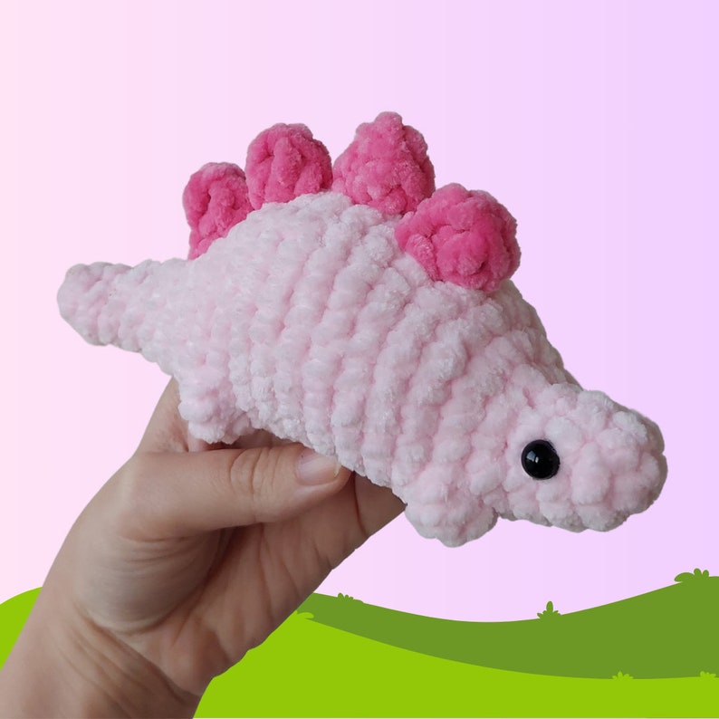 Puede incluir: Un juguete de dinosaurio de crochet rosa con p&uacute;as rosas en la espalda y ojos negros.