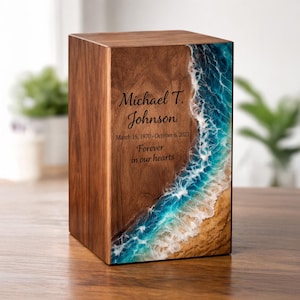 May include: A wooden urn with a decorative ocean wave design. The front of the urn features the name "Michael T. Johnson", birth and death dates, and the phrase "Forever in our hearts". The ocean design includes blue, white, and tan colors.