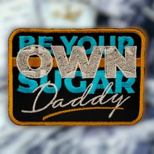 Könnte beinhalten: Ein schwarzer gestickter Aufnäher mit dem Text "Be Your Own Sugar Daddy" in weißer, blauer und goldener Farbe. Der Text "Own" ist in weiß und größer als der restliche Text.