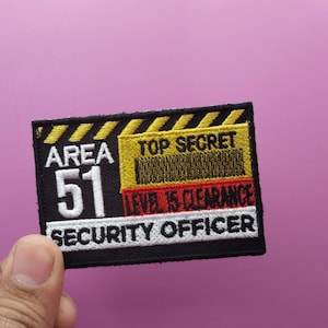Puede incluir: Parche negro bordado con rayas amarillas y rojas. El parche dice "Area 51" en texto blanco, "Top Secret" en texto amarillo, "Level 15 Clearance" en texto rojo y "Security Officer" en texto blanco.