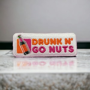 Pode incluir: Etiqueta branca bordada com texto rosa e laranja que diz "DRUNK N' GO NUTS". Uma pequena imagem de uma garrafa rosa e laranja está à esquerda do texto.