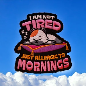 Puede incluir: Parche bordado con un diseño rosa y negro. Presenta un gato de dibujos animados durmiendo en una almohada morada con el texto "No estoy cansado, solo alérgico a las mañanas".