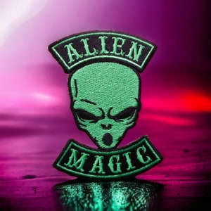 Può includere: Testa di alieno verde con le parole "Alien Magic" in ricamo nero su una toppa nera.