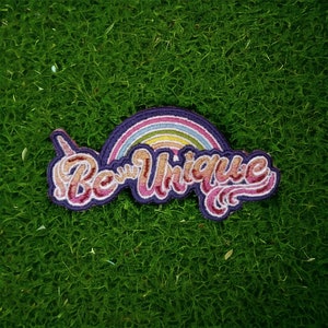Puede incluir: Un parche bordado colorido con un arcoíris y el texto "Be Unique" en una fuente retro.