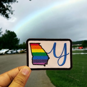 Könnte beinhalten: Ein Patch mit einem regenbogenfarbenen Umriss des Bundesstaates Georgia und dem Buchstaben "Y" in blauer Kursivschrift auf weißem Hintergrund. Der Patch wird in einer Hand vor einem Hintergrund aus einem Regenbogen und einem grünen Grasfeld gehalten.